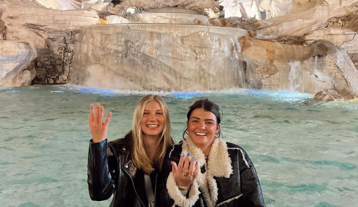 Two students sitting on the edge of the Trevi Fountain at night laughing, throwing coins behind them.