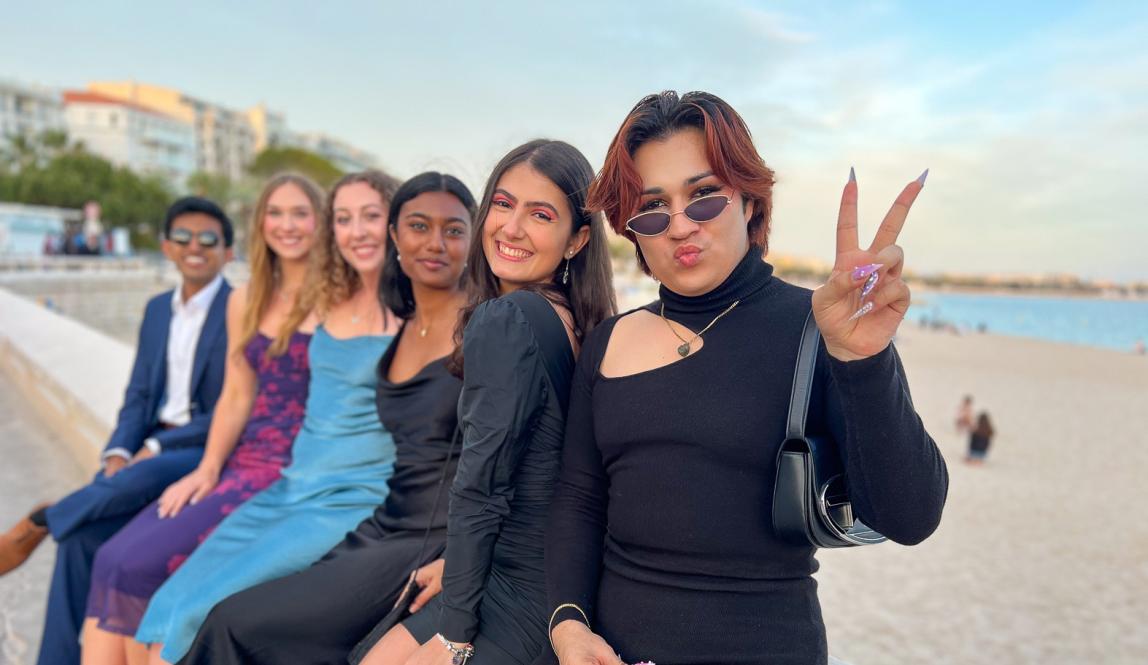 group of students dressed up fancy sitting on a bench with sand and water in the background