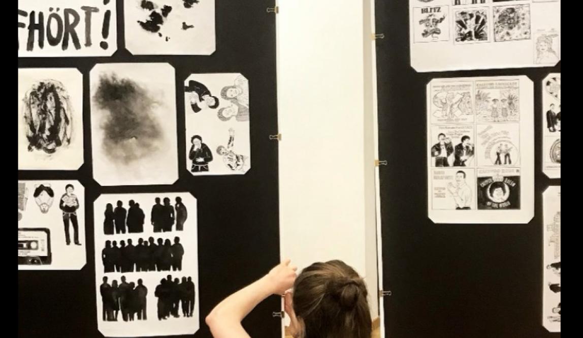 Young woman sitting on floor cross-legged, facing away from camera, securing original drawings to gallery wall 