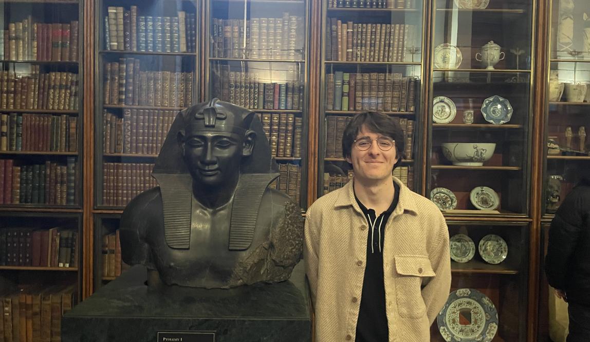 student standing with arms behind back next to a bust of a figure with books in the background behind glass doors