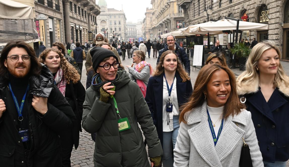 A very of several warmly dressed students and a professor with happy expressions on their faces as they walk through the streets of milan on a grey day