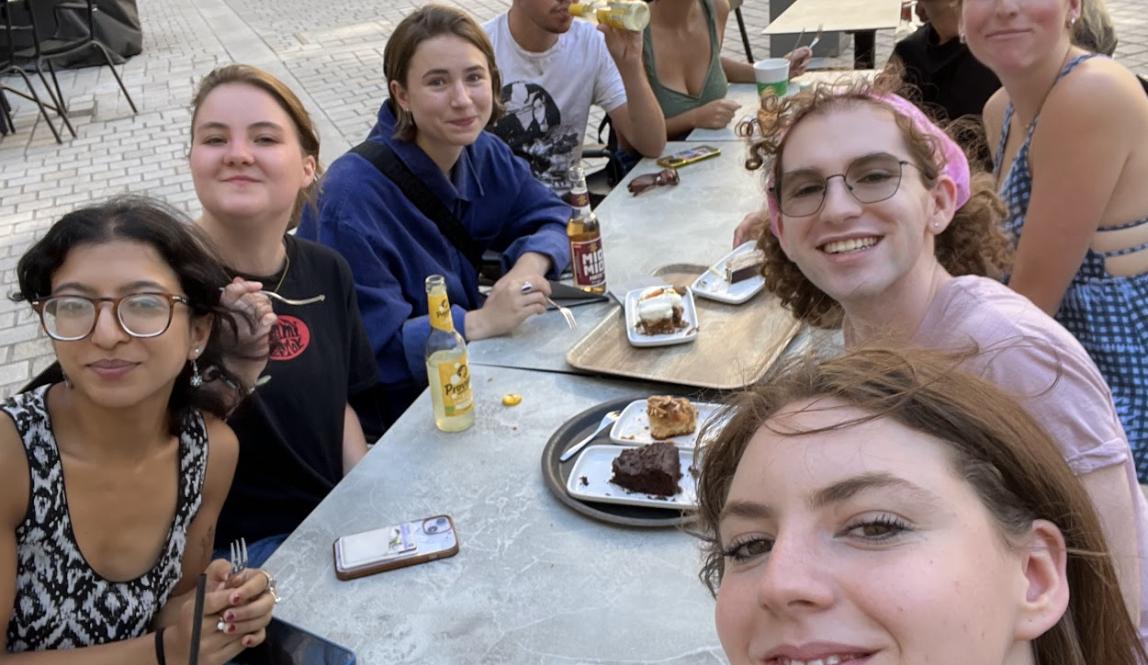 Selfie of a large group of students sitting outdoors at a long, white table 