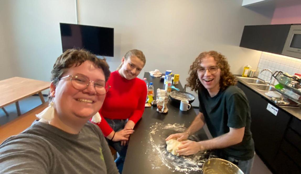 Selfie of three students around a cluttered counter in a communal kitchen. A space has been cleared on the counter, where one of the students is kneading dough.