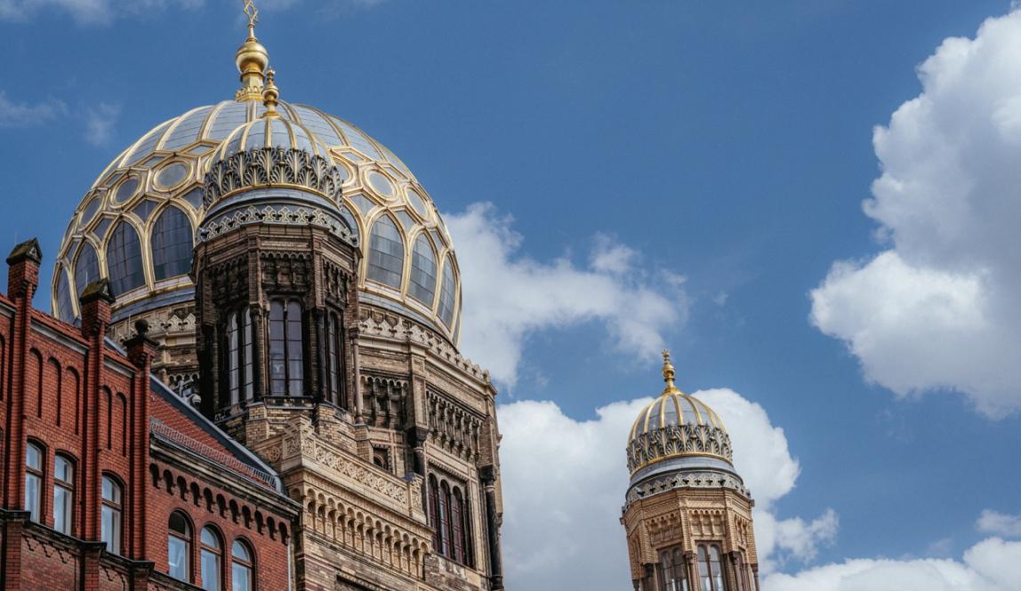 Photograph of the elaborately decorated dome of the Neues Synagogue in Berlin, with a cloudy, blue sky in the background. 