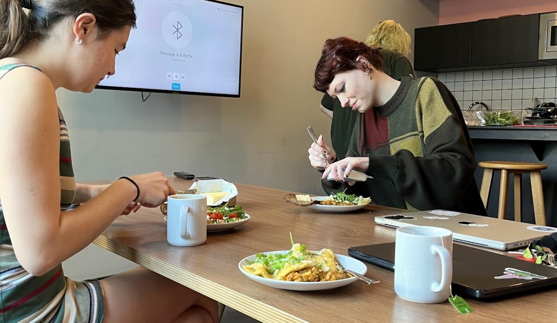 Candid photo of two students eating lunch and drinking at a kitchen table; another student can be seen in the background, preparing food at a counter. 