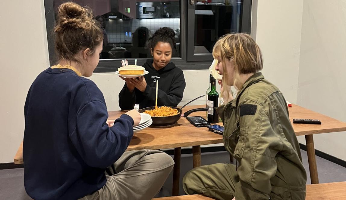 Three students sit at a kitchen table that is set out for a birthday meal. One student holds up a cake. 