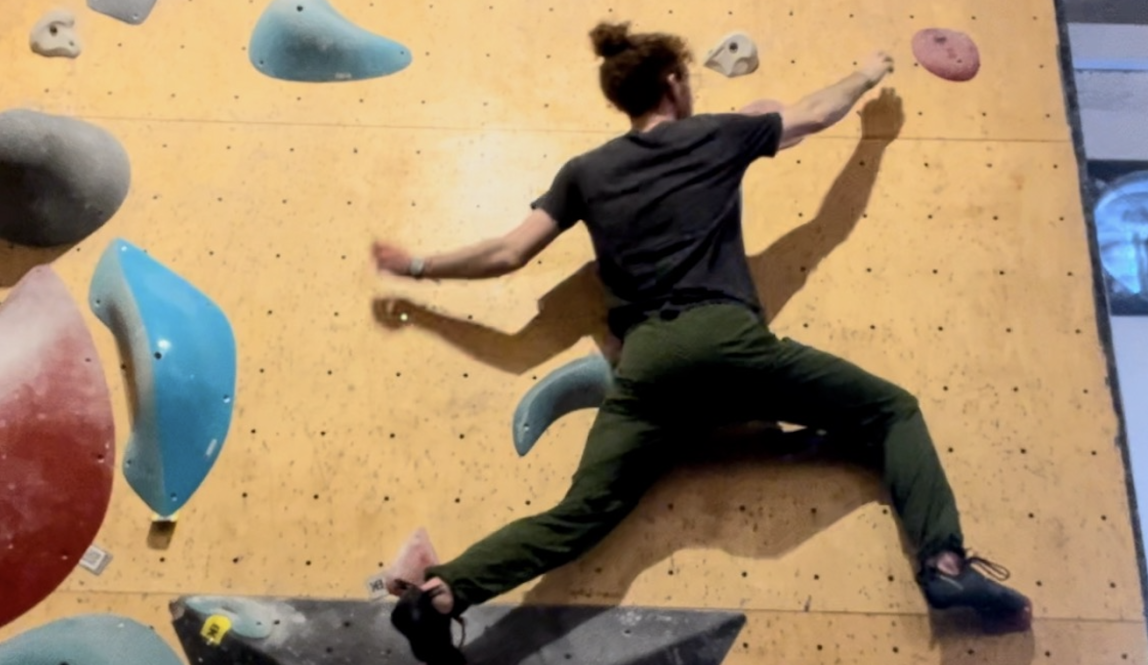 Photo of Tal on a bouldering wall, jumping horizontally to reach the next climbing hold. 