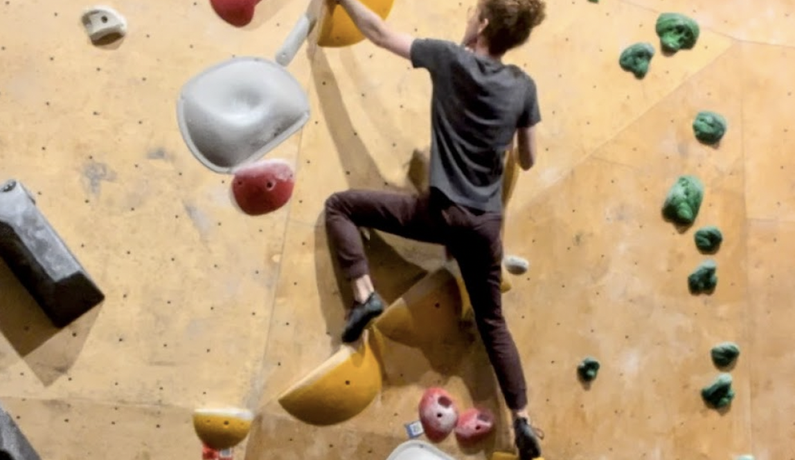 Photo of Tal on a bouldering wall, looking upwards at the final hold. 