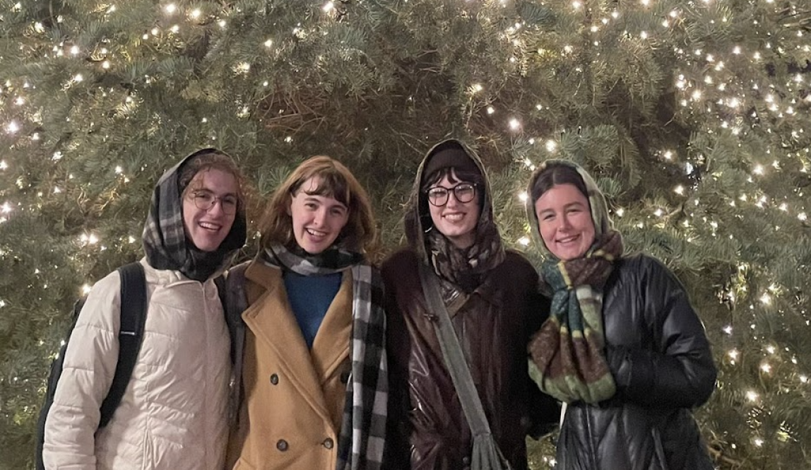 Four students pose in front of a large Christmas tree, bundled up in winter clothes. 