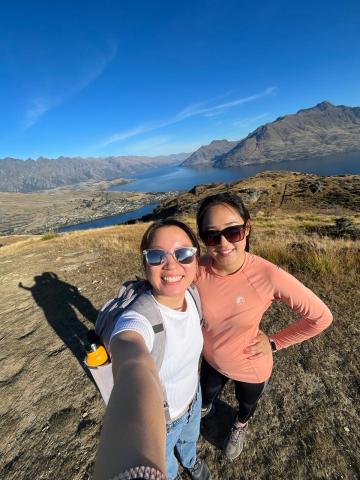 Two girls pose for a selfie at the top of the Queenstown Hill. The girl on the left wears a white shirt and jeans, and the girl on the right wears a pink shirt and black pants. The background shows a few mountains and a clear sky. 