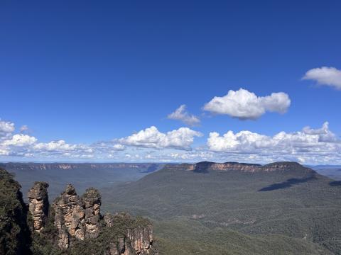 Three tan rock formations jut from the ground in the bottom left corner. The rest of the photo is forested land with a few mountains in the far distance. The sky is blue with bright white clouds.