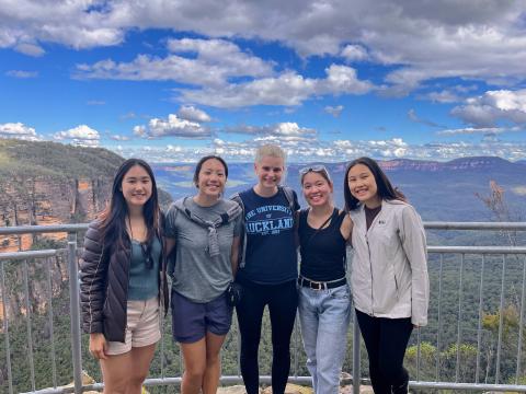 Five girls pose in front of a mountainous backdrop. The sky is filled with wispy and puffy clouds.