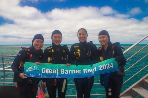 Four girls pose in blue stinger suits and hold a large banner that says Great Barrier Reef 2024.