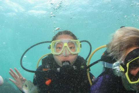 A girl waves at the camera as she scuba dives. She has goggles on her face and a mouthpiece helping her breathe. There is a fish in the bottom left corner.