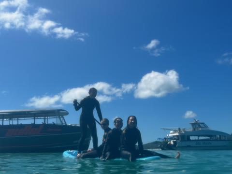 Three girls sit on a paddleboard while another girl stands up and paddles. Two boats are behind them, and the sky is clear with a few puffy clouds.