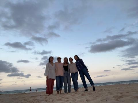 Five girls pose on a beach. The sky is orange blue as sunset is passing and there are dark clouds looming.