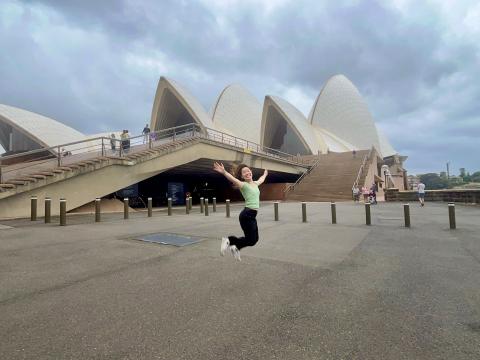 A girl wearing a green shirt and black pants jumps in front of the Sydney Opera House, a performing arts center that has distinctive pointed shell-like features as its roof.