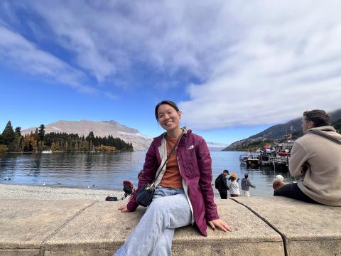 A girl sits atop a concrete structure and wears a purple rain jacket, jeans, and an orange shirt. She sits in front of the Queenstown landscape, consisting of water, trees, and cliffed regions.
