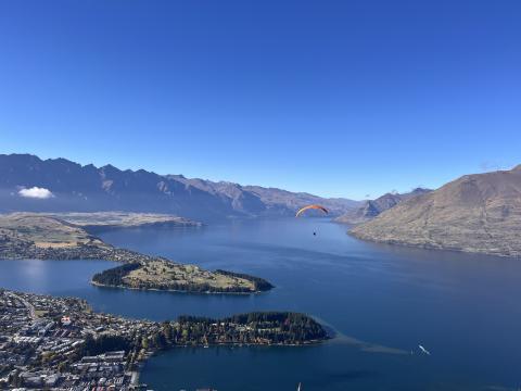 A vast body of water surrounded by several mountains and the buildings of Queenstown. A paraglider is shown in flight. 