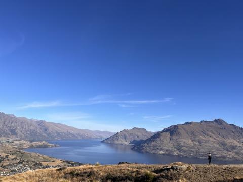 Landscape of Queenstown with a few mountains, vast water, and a clear sky. There is a small person in the right corner, showing the expanse of the landscape.