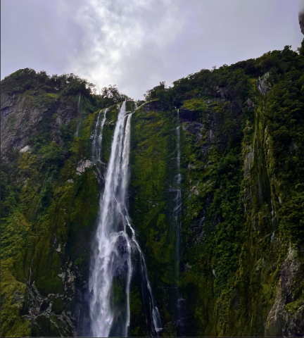 A large waterfall falls from a steep forested cliff. The sky is dark and cloudy.