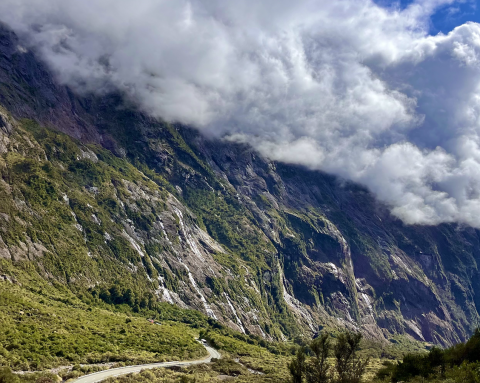 White clouds loom over the large mountain face of Milford Sound. There is a road that snakes through the landscape.