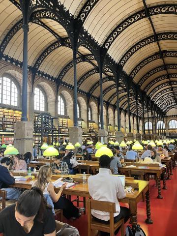 inside a library with tall ceilings and big arches; students are working at long wooden tables with lamps