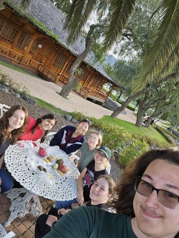 A selfie of seven students seated around an ornate metal table. Several different fruits sit atop the table.