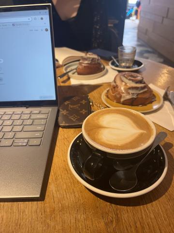a photo of a table in a café; to the left there is an open laptop and to the left is a hot cappuccino and a cinnamon roll