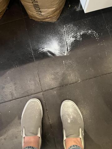 a photo of the floor; the floor is black and someone is wearing grey Vans shoes, which are visible in the photo; there are flour on both the floor and the shoes