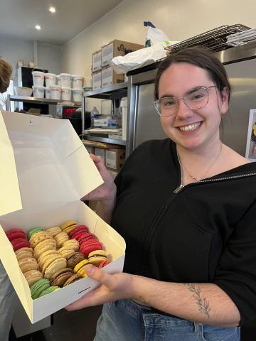 a photo inside a bakery kitchen; a girl is to the right and is holding a white box full of different types of macarons that she and her friends made for the first time