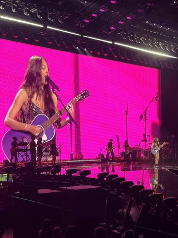 a photo of a stage in a large arena; Olivia Rodrigo is visible on screen and on the stage while playing the guitar