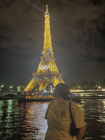 it is dark outside; the Eiffel Tower is in view across the Seine river and is sparkling; a girl stands and is admiring the tower with her head turned