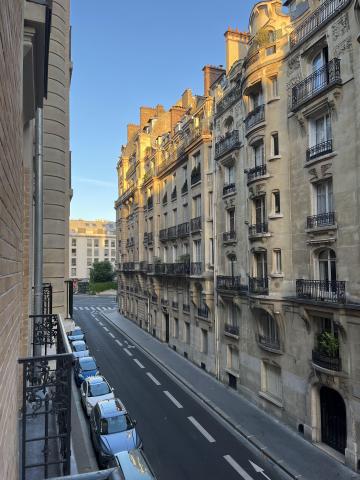 the photo is taken from the balcony of a residential building in Paris; it is early in the morning and the sun is rising, the sky is blue; there is a view down the street that included views of the buildings on the road