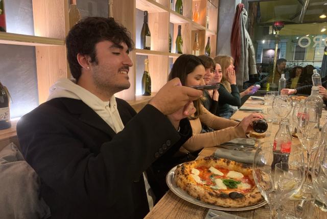 A male student is sitting at a table in a restaurant, smiling as he takes an arial photo of his pizza before eating it.