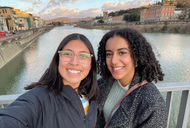 Two students are smiling while taking a selfie with the river in Rome behind them