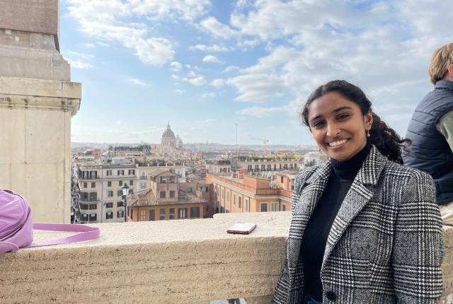 A student in a grey houndstooth blazer smiles from a terrace with the city of Rome sprawling behind her.