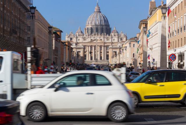 A timelapse photo of a busy street in front of the Vatican in Rome, with cards driving in front of the iconic building.