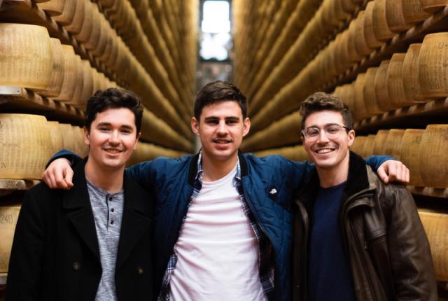 Three friends stand in front of a wall of aging cheese wheels, smiling.