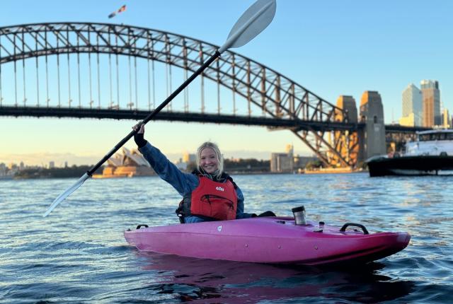 a student posing from a kayak in Sydney Harbour, holding their paddle in the air above their head in celebration
