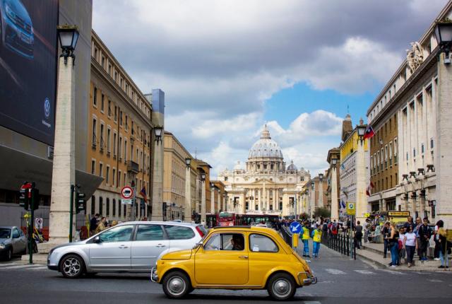 A vintage yellow car passes by on a busy street in front of the Vatican.