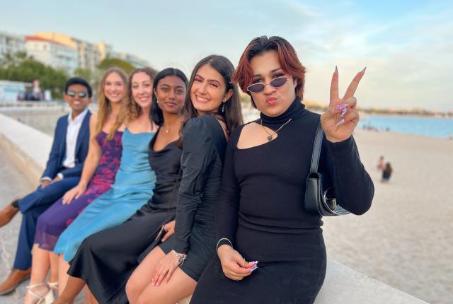 group of students dressed up fancy sitting on a bench with sand and water in the background