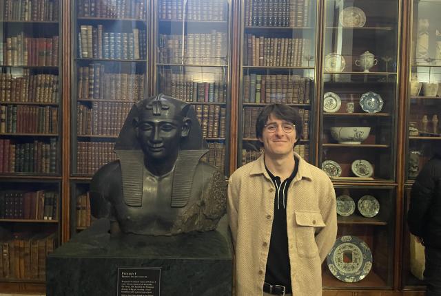 student standing with arms behind back next to a bust of a figure with books in the background behind glass doors