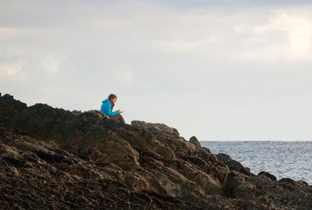 dublin student sitting on a rocky shore by the sea writing