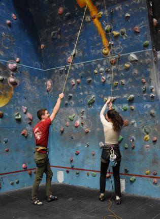 two dublin students standing in front of a rock climbing wall holding on to rope and smiling