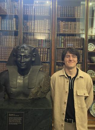 student standing with arms behind back next to a bust of a figure with books in the background behind glass doors