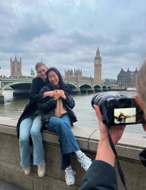 two students sitting on ledge hugging while another student holds up a camera and takes a picture