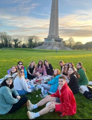 group of students sitting on the grass with picnic stuff in front of a stone monument