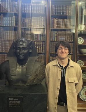 student standing with arms behind back next to a bust of a figure with books in the background behind glass doors