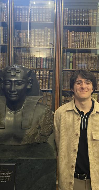 student standing with arms behind back next to a bust of a figure with books in the background behind glass doors